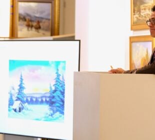 Aaron Bastian, Director of Fine Arts at Bonhams San Francisco, stands at an auctioneer’s podium in a brightly lit gallery, looking toward bidders while holding a pencil. A large screen beside him displays Bonhams auction details and an image of a Bob Ross winter landscape painting. Framed artworks hang on the walls behind him.