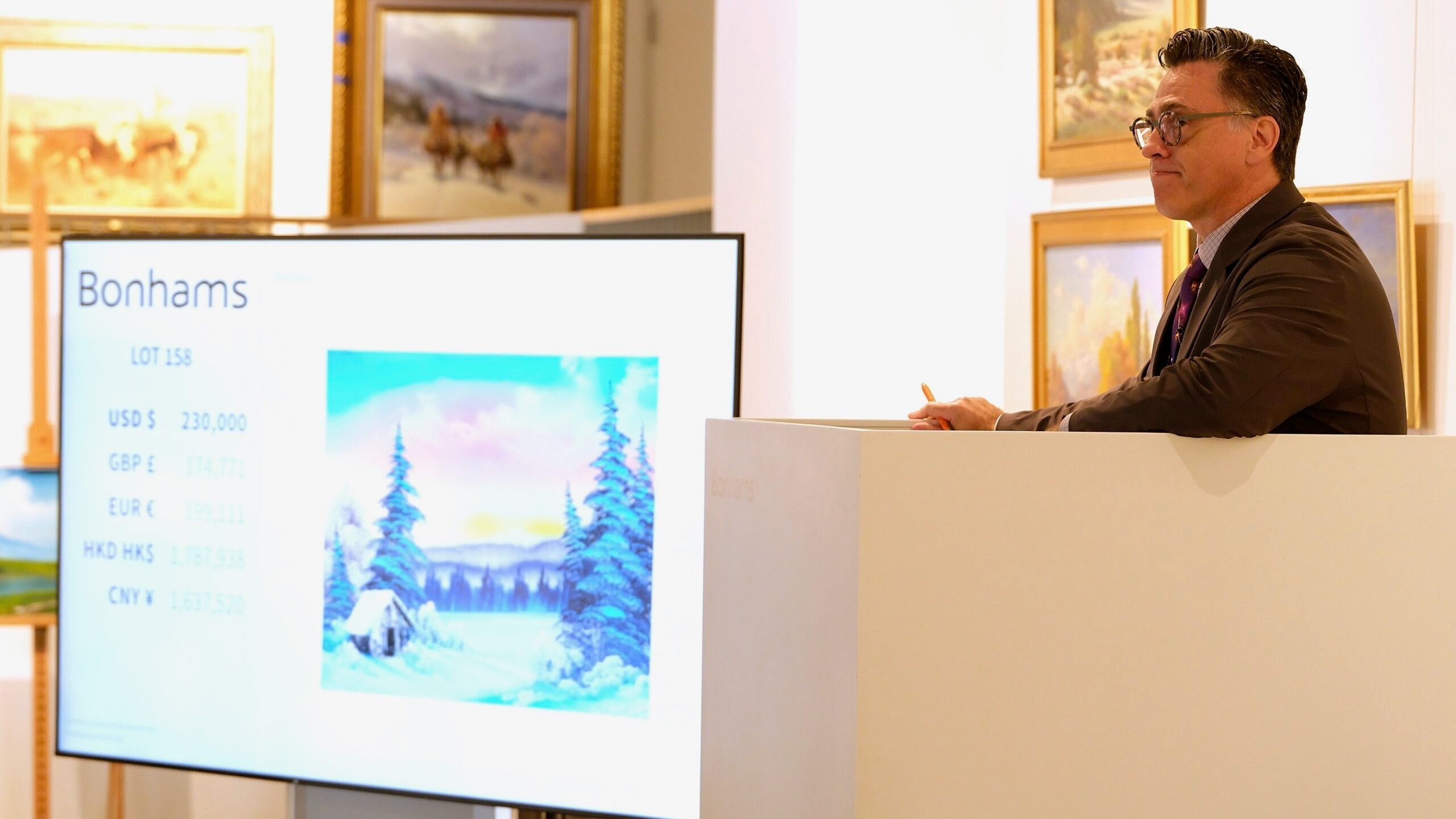 Aaron Bastian, Director of Fine Arts at Bonhams San Francisco, stands at an auctioneer’s podium in a brightly lit gallery, looking toward bidders while holding a pencil. A large screen beside him displays Bonhams auction details and an image of a Bob Ross winter landscape painting. Framed artworks hang on the walls behind him.