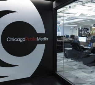 A view inside the Chicago Public Media office shows a large logo on the wall near glass windows looking into a modern workspace, where staff are working at desks with computers and boxes in the background.