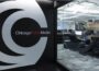 A view inside the Chicago Public Media office shows a large logo on the wall near glass windows looking into a modern workspace, where staff are working at desks with computers and boxes in the background.