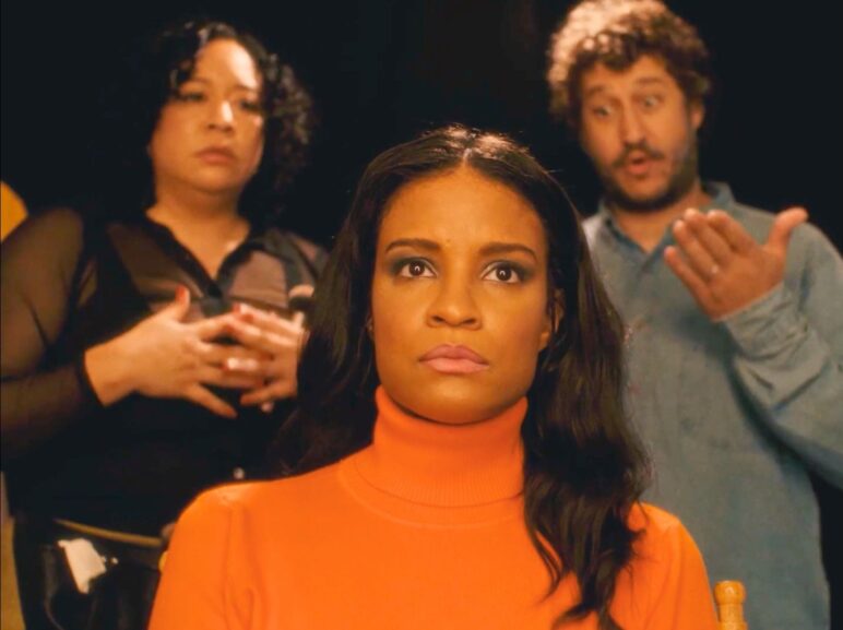 In a dramatized scene, a woman in an orange turtleneck sits in the foreground with a tense, focused expression. Behind her, a woman in a black sheer top holds her hands to her chest, looking concerned, while a man in a blue shirt gestures animatedly as if giving direction. The background is dark, drawing attention to the three performers.