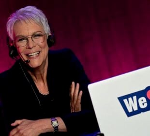 Jamie Lee Curtis sits at a desk wearing a headset microphone and glasses. She is dressed in a dark blazer and is illuminated by warm lighting against a deep magenta backdrop. A white laptop in front of her displays a “We” followed by a red heart symbol.
