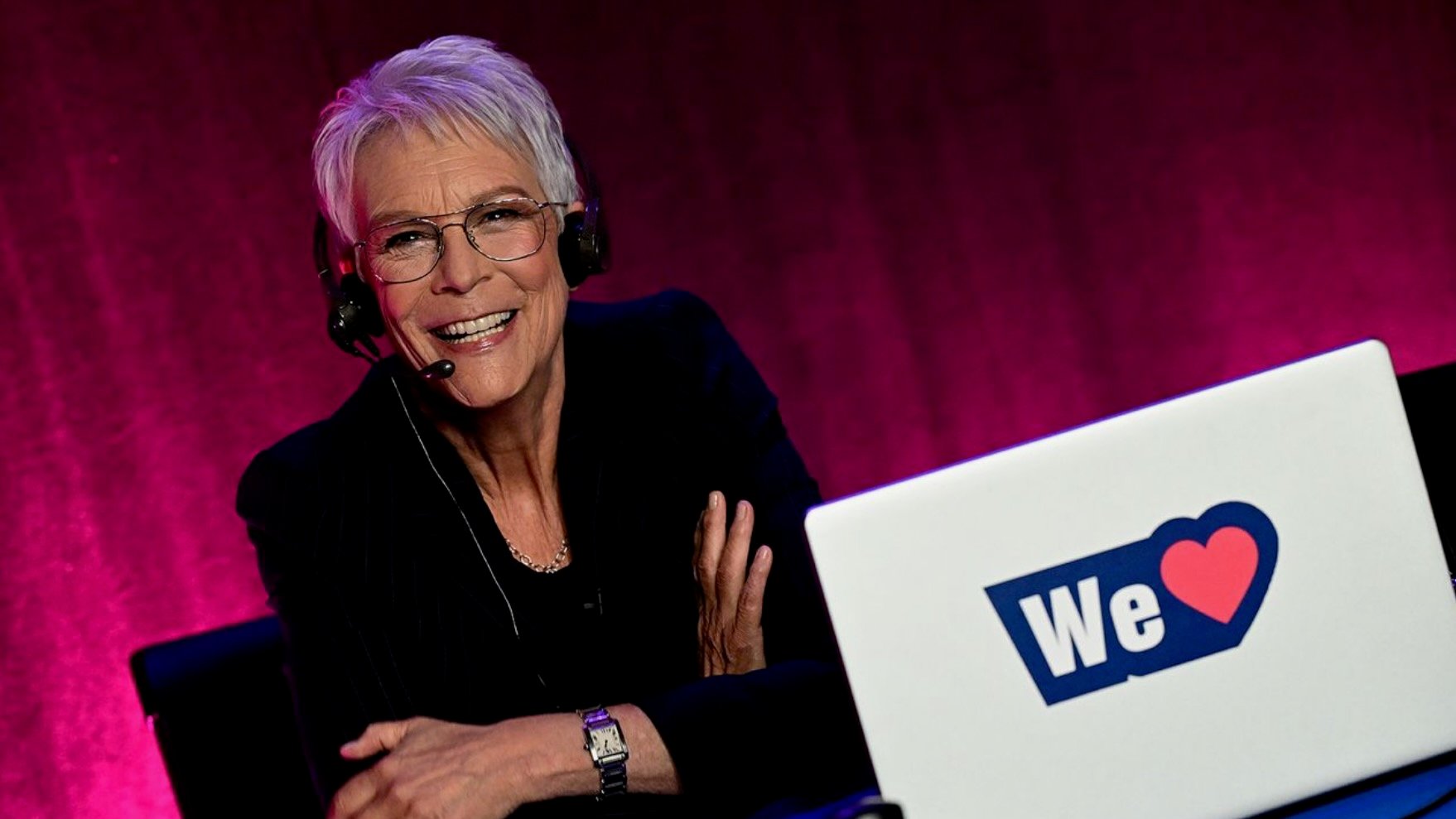 Jamie Lee Curtis sits at a desk wearing a headset microphone and glasses. She is dressed in a dark blazer and is illuminated by warm lighting against a deep magenta backdrop. A white laptop in front of her displays a “We” followed by a red heart symbol.