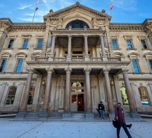 Wide-angle view of the New Jersey Statehouse’s ornate stone façade under a blue sky, with tall columns framing the main entrance. Two journalists walk away from the building on the front walkway, carrying equipment, after exiting through the open doorway.