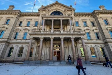 Wide-angle view of the New Jersey Statehouse’s ornate stone façade under a blue sky, with tall columns framing the main entrance. Two journalists walk away from the building on the front walkway, carrying equipment, after exiting through the open doorway.