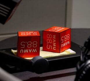Two red WAMU 88.5 microphone flags sit on a notepad on a studio table, surrounded by broadcast equipment including a microphone arm and a pair of headphones.