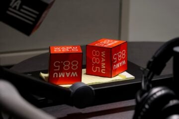 Two red WAMU 88.5 microphone flags sit on a notepad on a studio table, surrounded by broadcast equipment including a microphone arm and a pair of headphones.