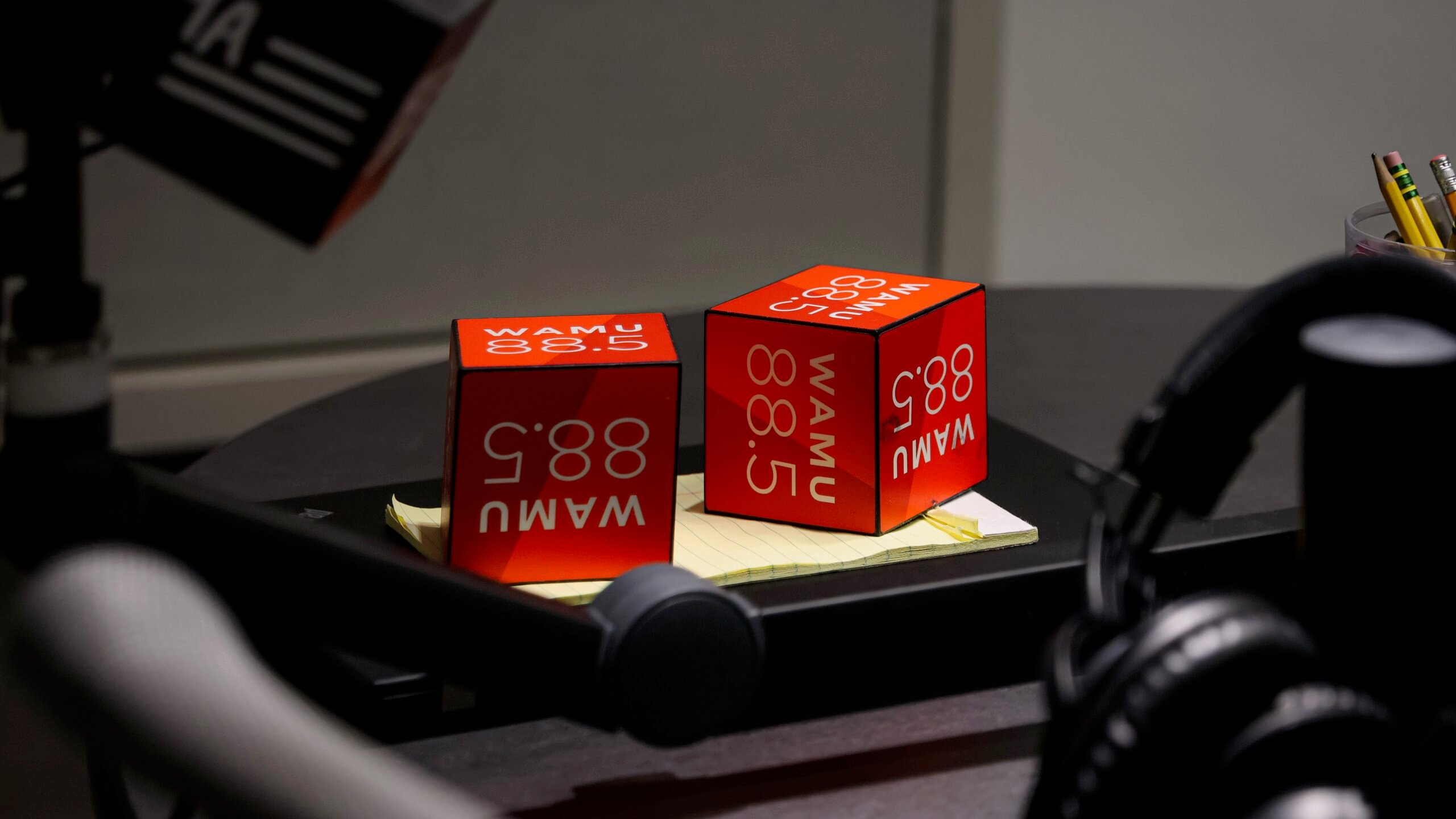 Two red WAMU 88.5 microphone flags sit on a notepad on a studio table, surrounded by broadcast equipment including a microphone arm and a pair of headphones.