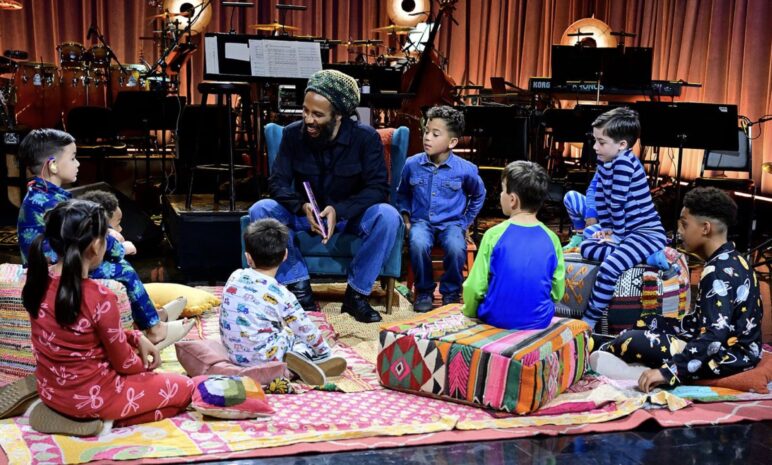 Musician Ziggy Marley sits on a chair holding a book while a group of young children in pajamas gather around him on colorful rugs and cushions. The children sit in a semicircle, listening attentively. Behind them is a stage setup with musical instruments, including drums, keyboards, and stands, softly lit by warm studio lights.