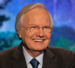 Bill Moyers smiles warmly while wearing a dark suit, blue shirt and tie, seated in front of a softly blurred, colorful background.
