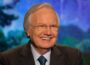 Bill Moyers smiles warmly while wearing a dark suit, blue shirt and tie, seated in front of a softly blurred, colorful background.
