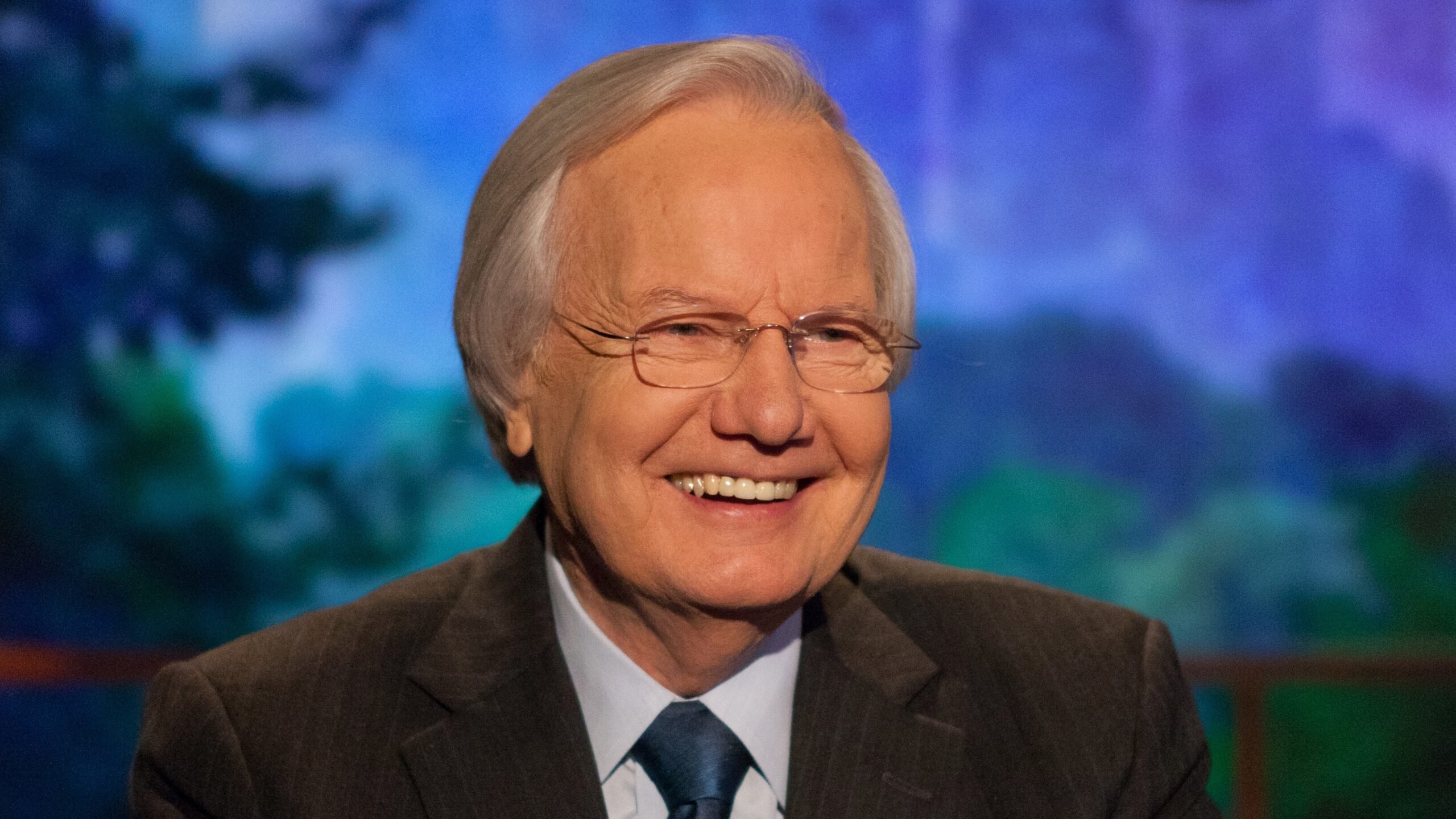 Bill Moyers smiles warmly while wearing a dark suit, blue shirt and tie, seated in front of a softly blurred, colorful background.