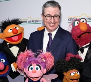 John Oliver smiles while posing on a red carpet with several Sesame Street Muppets dressed in formal tuxedos, standing in front of a step-and-repeat backdrop featuring the Sesame Workshop logo.