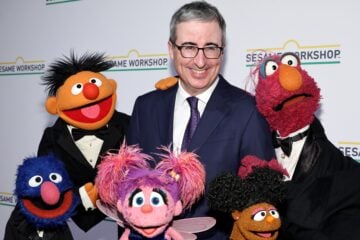 John Oliver smiles while posing on a red carpet with several Sesame Street Muppets dressed in formal tuxedos, standing in front of a step-and-repeat backdrop featuring the Sesame Workshop logo.