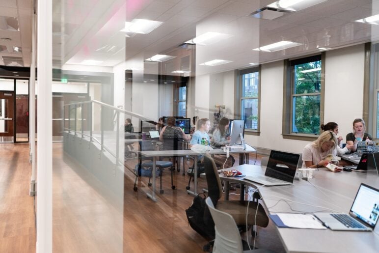 Students work at shared tables in the Marquette Wire news studio at Marquette University, using desktop computers and laptops. The space has glass walls, overhead fluorescent lighting and large windows along one side, with people focused on screens, talking quietly or eating snacks while working.
