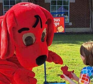 A person in a Clifford the Big Red Dog costume bends down toward a young child outdoors, reaching out as the child holds a small red dog toy. A brick building with windows and a sign for Curious George is visible in the background on a sunny day.