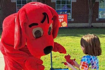 A person in a Clifford the Big Red Dog costume bends down toward a young child outdoors, reaching out as the child holds a small red dog toy. A brick building with windows and a sign for Curious George is visible in the background on a sunny day.