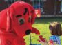 A person in a Clifford the Big Red Dog costume bends down toward a young child outdoors, reaching out as the child holds a small red dog toy. A brick building with windows and a sign for Curious George is visible in the background on a sunny day.