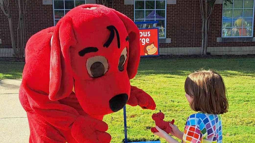 A person in a Clifford the Big Red Dog costume bends down toward a young child outdoors, reaching out as the child holds a small red dog toy. A brick building with windows and a sign for Curious George is visible in the background on a sunny day.
