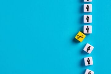 Line of small white blocks with simple human icons arranged vertically on a bright blue surface, with one yellow block showing scissors tilted out of line to suggest a job cut or layoff.