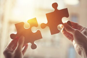 Two hands hold separate cardboard puzzle pieces up toward warm backlight, positioning them to fit together.