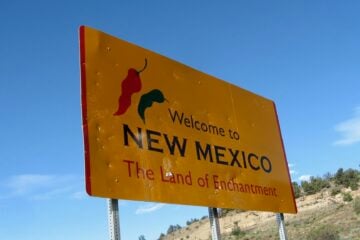 Yellow roadside sign reading “Welcome to New Mexico — The Land of Enchantment,” with red and green chili pepper graphics, set against a blue sky and desert hillside.