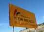 Yellow roadside sign reading “Welcome to New Mexico — The Land of Enchantment,” with red and green chili pepper graphics, set against a blue sky and desert hillside.