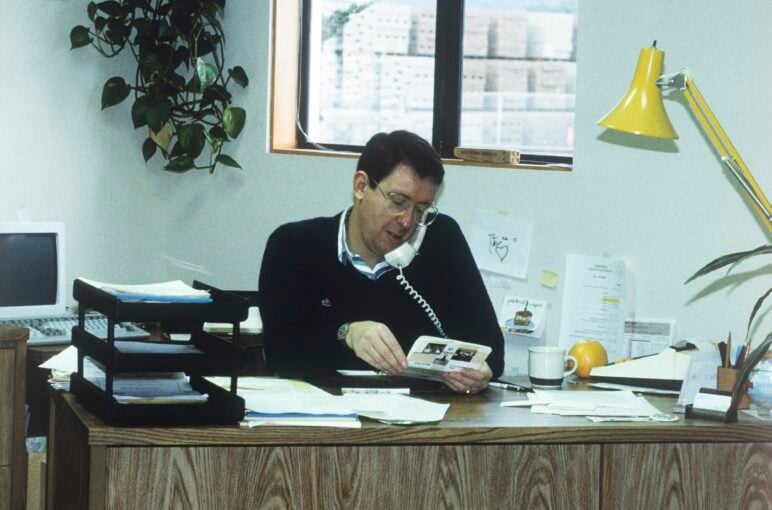 Frank Lanzone sits at an office desk, holding a telephone receiver to his shoulder; behind him are a window and a hanging plant, with stacks of documents, a computer monitor, office supplies and a yellow desk lamp arranged around the workspace.