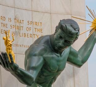 The Spirit of Detroit, a bronze statue of a male figure holding a radiant golden orb in one hand and smaller gilded human figures in the other, set against a stone wall engraved with a Bible verse.