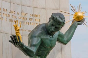 The Spirit of Detroit, a bronze statue of a male figure holding a radiant golden orb in one hand and smaller gilded human figures in the other, set against a stone wall engraved with a Bible verse.