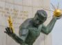 The Spirit of Detroit, a bronze statue of a male figure holding a radiant golden orb in one hand and smaller gilded human figures in the other, set against a stone wall engraved with a Bible verse.