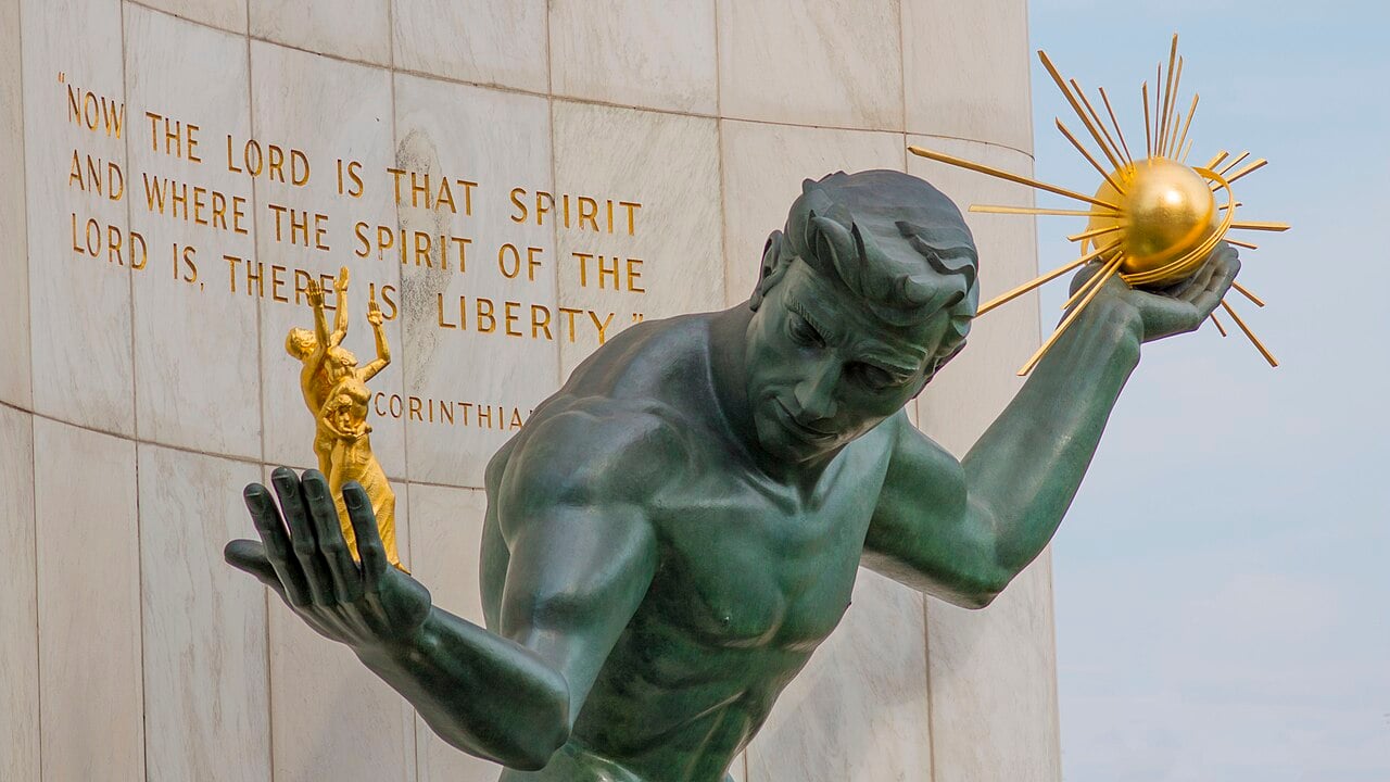The Spirit of Detroit, a bronze statue of a male figure holding a radiant golden orb in one hand and smaller gilded human figures in the other, set against a stone wall engraved with a Bible verse.