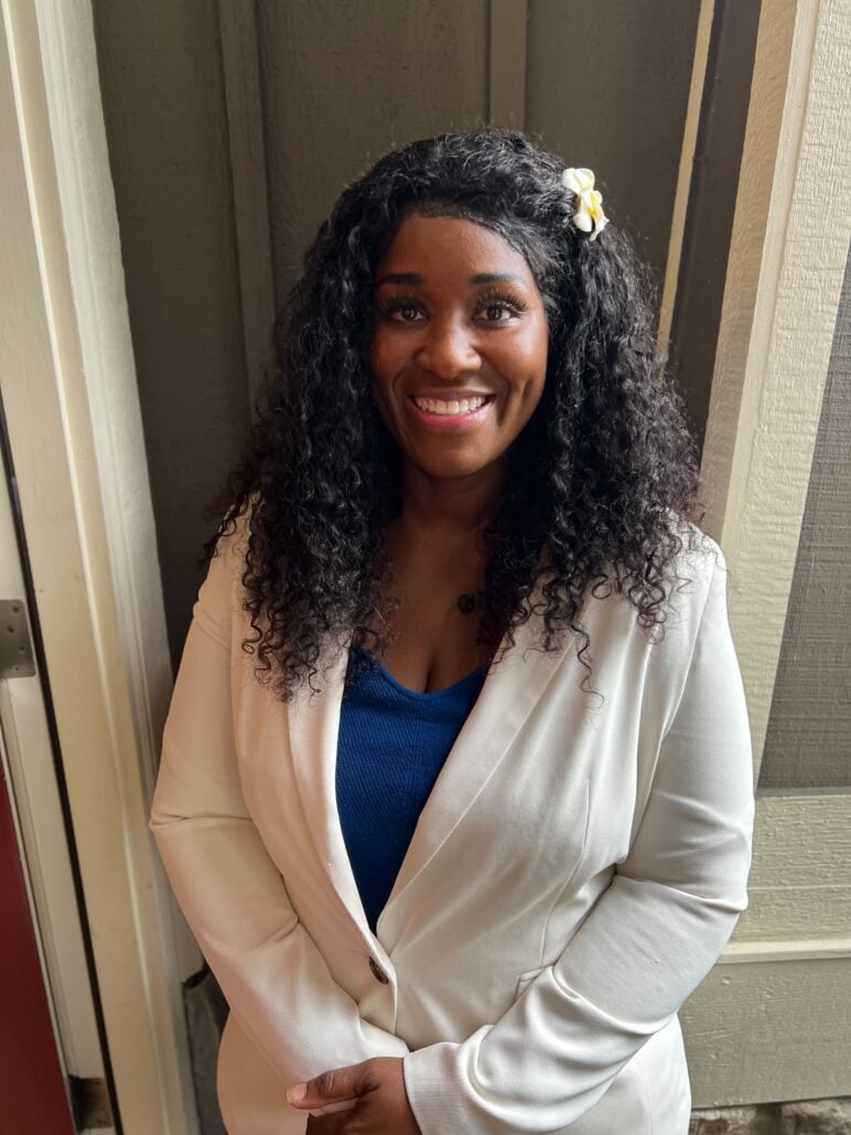 With long curly hair and a white flower clip, Fieta Robinson stands with her hands clasped, wearing a white blazer over a blue top, photographed in soft light near a doorway.