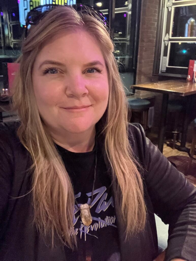 Jess Berg smiles at the camera, with long blond hair and sunglasses resting on her head.