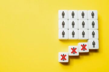 White cubes with stick-figure person icons are arranged in a grid on a bright yellow background. Several cubes have fallen away from the group, and three of them are marked with large red Xs, symbolizing layoffs or people being removed from a workforce