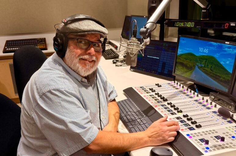 KVMR GM Luis Estrada sits at a mixing board in a studio with a microphone in front of him. He is wearing headphones and a gray hat. A computer monitor displays the time and a scene of blue skies over a mountaintop.