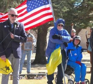 Wyoming Public Radio listener Richard Garrett speaks into a microphone at an outdoor rally while people stand nearby holding an American flag.