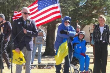 Wyoming Public Radio listener Richard Garrett speaks into a microphone at an outdoor rally while people stand nearby holding an American flag.