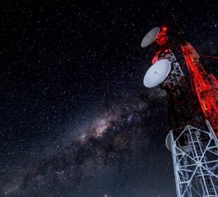 A tall communications tower with several dish antennas rises into a clear night sky filled with stars and the Milky Way, illuminated by red warning lights.