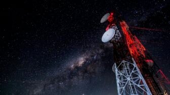 A tall communications tower with several dish antennas rises into a clear night sky filled with stars and the Milky Way, illuminated by red warning lights.