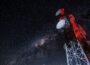 A tall communications tower with several dish antennas rises into a clear night sky filled with stars and the Milky Way, illuminated by red warning lights.