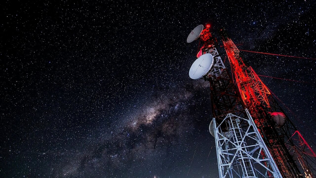 A tall communications tower with several dish antennas rises into a clear night sky filled with stars and the Milky Way, illuminated by red warning lights.