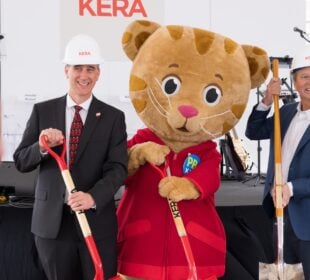 Wearing white KERA-branded hard hats, KERA CEO Nico Leone and Bill Addy, co-founder of the Addy Foundation, join Daniel Tiger holding ceremonial shovels during the groundbreaking event for KERA's new headquarters, with microphones, instruments and additional shovels visible behind them.