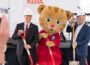 Wearing white KERA-branded hard hats, KERA CEO Nico Leone and Bill Addy, co-founder of the Addy Foundation, join Daniel Tiger holding ceremonial shovels during the groundbreaking event for KERA's new headquarters, with microphones, instruments and additional shovels visible behind them.