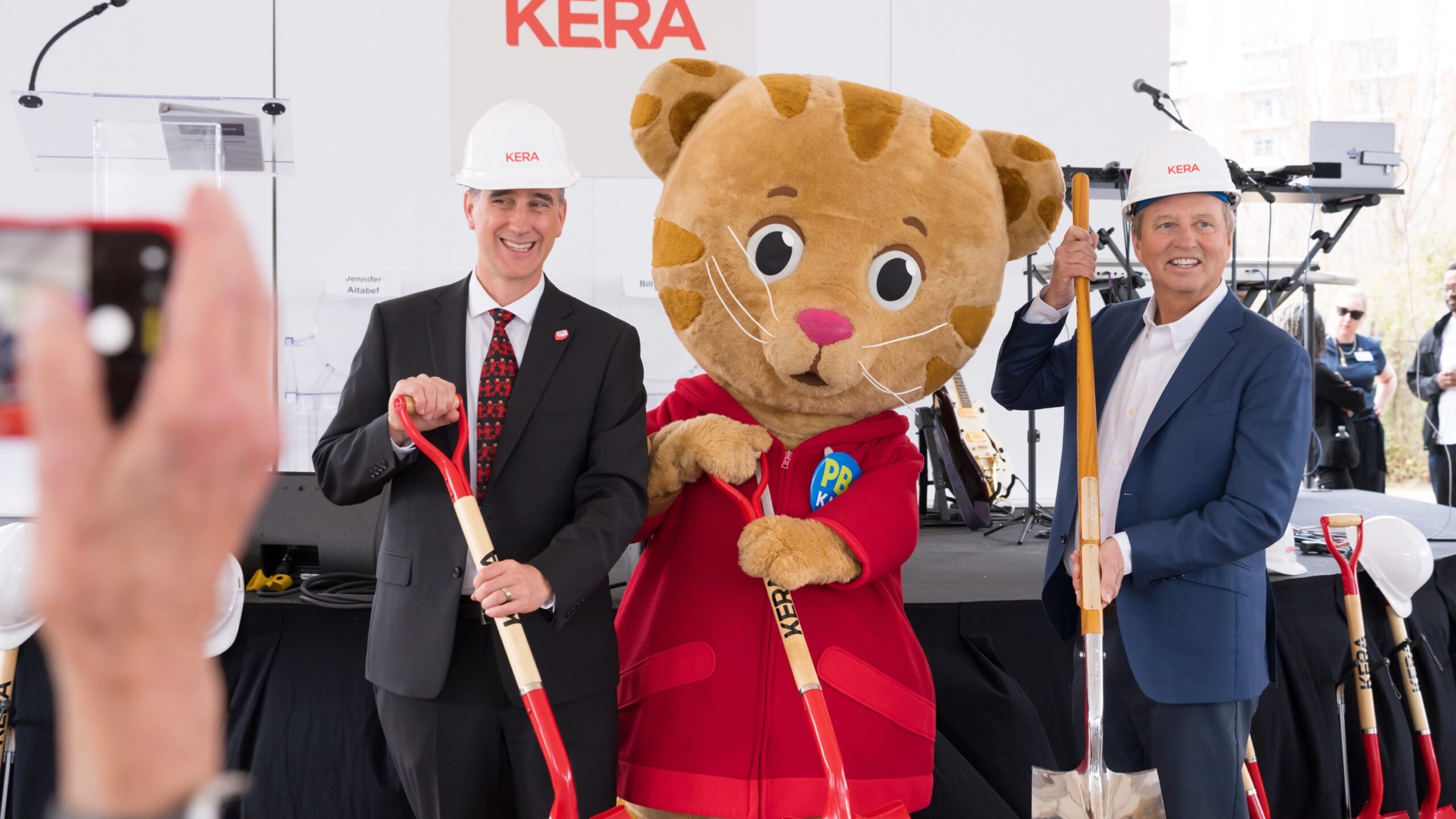 Wearing white KERA-branded hard hats, KERA CEO Nico Leone and Bill Addy, co-founder of the Addy Foundation, join Daniel Tiger holding ceremonial shovels during the groundbreaking event for KERA's new headquarters, with microphones, instruments and additional shovels visible behind them.