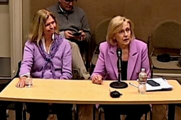 Nashville PBS CEO Becky Magura and PBS CEO Paula Kerger sit at a table during a public meeting. Kerger, wearing a pink blazer, speaks into a microphone while papers, a phone and a water bottle sit in front of her. Magura, in a purple sweater, sits beside her listening, while a man behind them looks down at a phone. Chairs and meeting-room equipment are visible in the background.