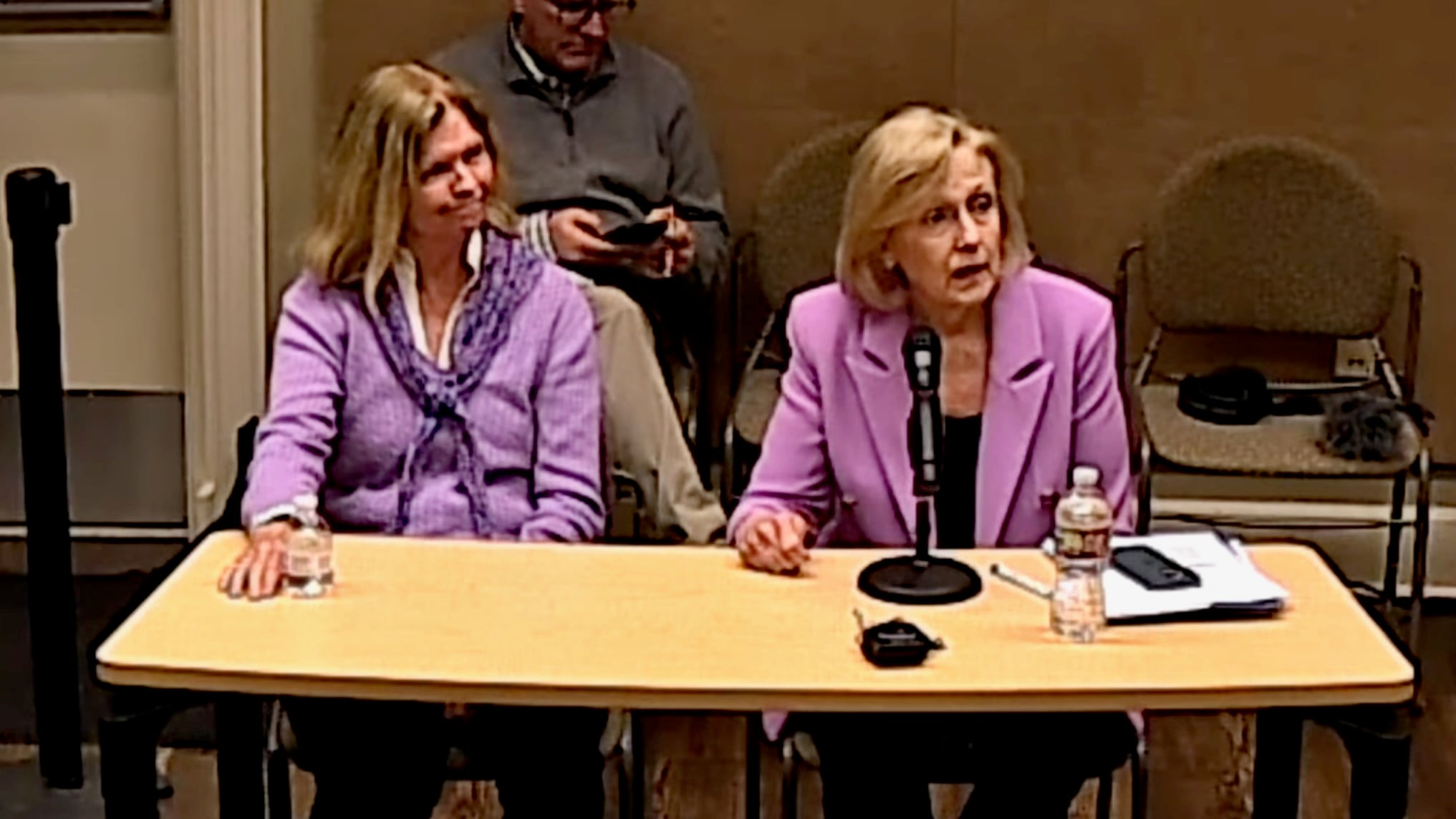 Nashville PBS CEO Becky Magura and PBS CEO Paula Kerger sit at a table during a public meeting. Kerger, wearing a pink blazer, speaks into a microphone while papers, a phone and a water bottle sit in front of her. Magura, in a purple sweater, sits beside her listening, while a man behind them looks down at a phone. Chairs and meeting-room equipment are visible in the background.