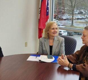 Former Arkansas First Lady Gay White, widow of Gov. Frank White; PBS CEO Paula Kerger; and former Arkansas First Lady Barbara Pryor, widow of Gov. David Pryor sit around a conference table in an office with large windows overlooking a parking lot on a rainy day. A folder and phone lie on the table, and an Arkansas state flag stands behind them.