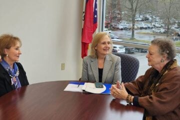Former Arkansas First Lady Gay White, widow of Gov. Frank White; PBS CEO Paula Kerger; and former Arkansas First Lady Barbara Pryor, widow of Gov. David Pryor sit around a conference table in an office with large windows overlooking a parking lot on a rainy day. A folder and phone lie on the table, and an Arkansas state flag stands behind them.