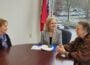 Former Arkansas First Lady Gay White, widow of Gov. Frank White; PBS CEO Paula Kerger; and former Arkansas First Lady Barbara Pryor, widow of Gov. David Pryor sit around a conference table in an office with large windows overlooking a parking lot on a rainy day. A folder and phone lie on the table, and an Arkansas state flag stands behind them.